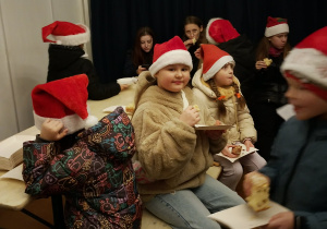 Uczniowie SP 13 w czapkach Mikołaja jedzą świąteczny poczęstunek przy ławkach w namiocie jarmarku.