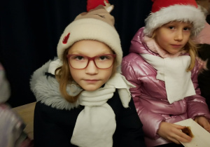 Dwie dziewczynki w zimowych kurtkach, białych szalikach i świątecznych czapkach siedzą obok siebie, w tle widać innych uczestników jarmarku.