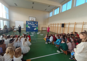 Uczniowie klas 1–3 w sali gimnastycznej podczas uroczystości, pani dyrektor w czerwonej sukience przemawia do dzieci.