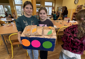 Uczennice w świetlicy szkolnej trzymają własnoręcznie wykonany kartonowy karmnik dla ptaków podczas zajęć plastycznych.