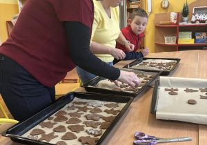 Dzieci w świetlicy szkolnej przygotowują pierniki, układając świąteczne kształty na blachach do pieczenia pod opieką nauczycieli.