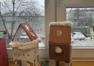 Dwa ręcznie wykonane karmniki dla ptaków z kartonu, ozdobione farbami i watą, prezentowane w świetlicy szkolnej.