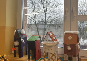 Kartonowe karmniki dla ptaków wykonane przez dzieci, ustawione na parapecie w świetlicy szkolnej.