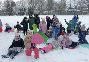 Uczniowie klasy 2b bawią się na śniegu, trzymając jabłuszka i sanki podczas zimowego wyjścia.