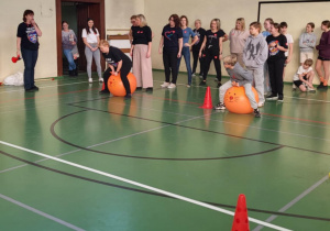 Uczniowie i nauczyciele rywalizują w sportowej konkurencji w sali gimnastycznej, pokonując tor z pachołkami na piłkach do skakania.