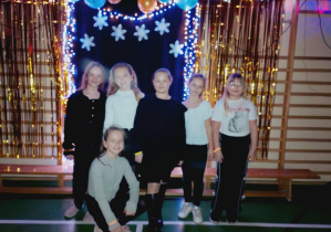 Sześć uczennic pozuje na tle dekoracji z balonami, śnieżynkami i światełkami podczas szkolnej dyskoteki karnawałowej. Dziewczynki ubrane w dyskotekowe stroje, uśmiechają się do zdjęcia w udekorowanej sali gimnastycznej.