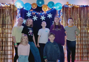 Uczniowie klasy piątej pozują podczas szkolnej dyskoteki karnawałowej na tle ozdób: balonów, śnieżynek i lampek. Grupa składa się z siedmiorga dzieci stojących na ławeczce i przed nią w sali gimnastycznej.