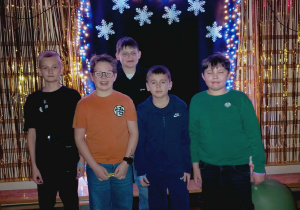 Grupa pięciu uczniów klasy czwartej pozuje na tle kolorowej dekoracji karnawałowej z balonami i światełkami.